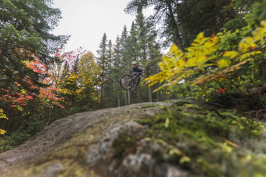 A mountain biker performing a jump over a rock in a forested area, surrounded by trees displaying autumn foliage. The image captures the dynamic movement of the rider in mid-air, with a focus on the natural scenery and vibrant colors of the leaves. Sentiers Du Moulin mountain bike trail.