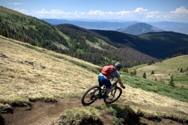 A mountain biker navigates a winding dirt trail through a grassy, mountainous landscape, with lush green trees and distant blue mountains under a partly cloudy sky.