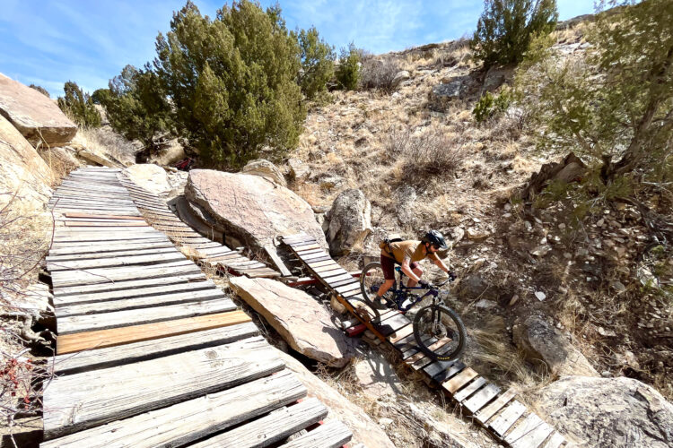 A mountain biker navigating a narrow, wooden footbridge surrounded by rocky terrain and sparse vegetation on a sunny day. The path is elevated over a rugged landscape, with large boulders and dry grass in the background.