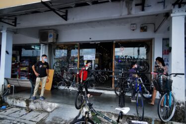 Diamondback Response Sport: A bicycle store named "Khass" with a large sign in front. The entrance is visible, showcasing a variety of bicycles on display. Two customers are interacting near the entrance, while several bicycles are parked outside on the pavement. The scene is urban, with a concrete walkway leading up to the shop.