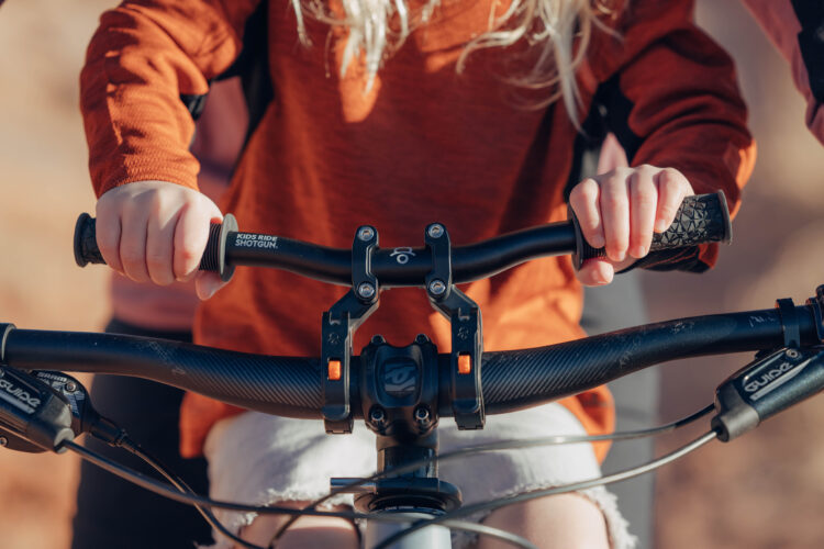 Close-up of a child’s hands gripping the handlebars of a mountain bike, wearing an orange long-sleeve shirt. The bike features a black grip and handlebar with a visible brand name "KIDS RIDE SHOTGUN." In the background, there is a blurry view of another person, suggesting a biking activity. The setting appears to be outdoors with a warm, sunny atmosphere.