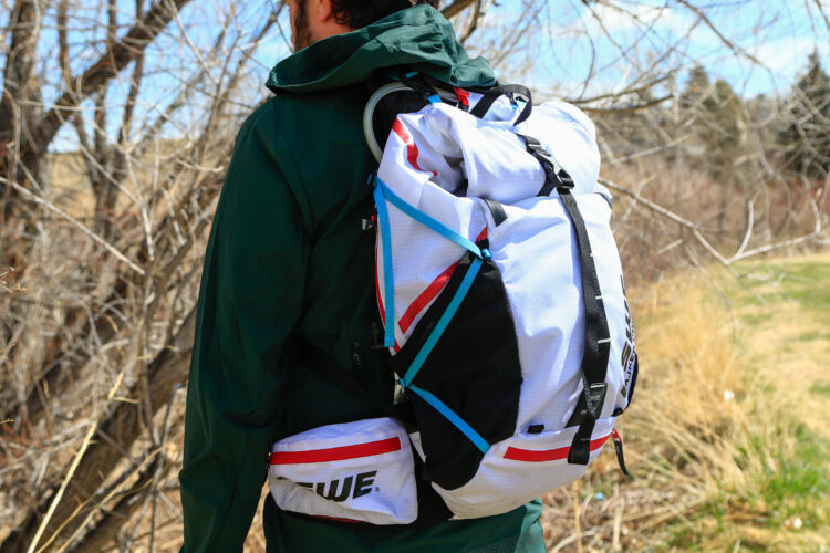 A person standing outdoors, wearing a green jacket and a multi-colored backpack, featuring white, black, and red elements. The backpack is secured with straps and includes an additional lower pouch. Bare trees and grass are visible in the background, indicating a natural setting.