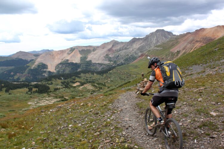 A mountain biker rides along a rugged trail with scenic views of mountains and valleys in the background. The cyclist, wearing a helmet and backpack, explores a lush, green landscape under a partly cloudy sky.
