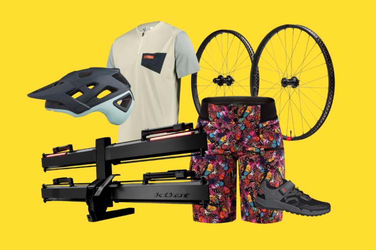 A collection of biking gear on a vibrant yellow background, featuring a blue cycling helmet, a light-colored cycling shirt, colorful patterned shorts, a pair of black cycling shoes, a bike rack, and two bicycle wheels.