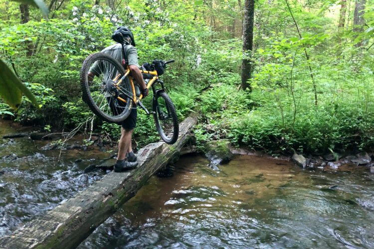 A mountain biker balancing on a wooden log bridge over a stream while carrying their bike, surrounded by lush green foliage in a forest setting.