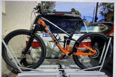 A mountain bike with an orange and gray frame mounted on a bike rack attached to an SUV. The bike has thick tires and is secured in place, while the background shows a partially cloudy sky and various vehicles parked nearby.