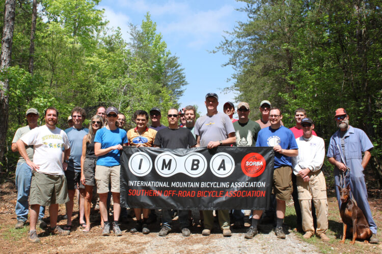 A group photo of individuals standing together in a wooded area, holding a banner for the International Mountain Bicycling Association (IMBA) and the Southern Off-Road Bicycle Association (SORBA). The group consists of diverse individuals, including men and women dressed casually, with some wearing hats and sunglasses. There is a dog on a leash in the foreground. The scene is bright and sunny, surrounded by lush green trees.