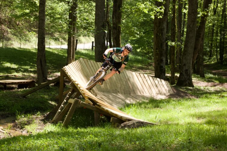 A mountain biker riding on a wooden ramp through a forested area, leaning into the turn with a focused expression. The scene features lush green grass and trees, creating a vibrant outdoor setting for biking.