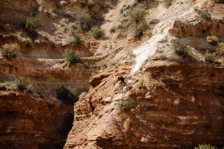 A mountain biker performing a daring jump off a rocky cliff in a rugged, arid landscape, with dust and debris flying from the impact on the ground below.