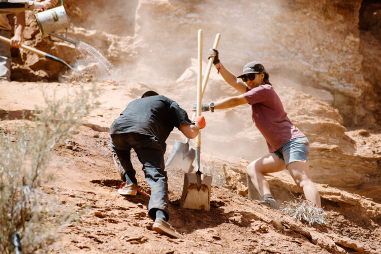 Two individuals are engaged in outdoor manual labor on a rocky terrain, with dust rising around them. One person, wearing a purple shirt and sunglasses, stands on a slope using a shovel, while the other, dressed in a black shirt and gloves, crouches down with a shovel and seems to be digging. The background features rocky formations, and there are additional people working in the distance, adding to the scene of teamwork and effort.