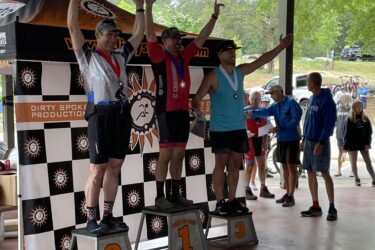 Three male athletes stand on podiums, celebrating after a competition, with arms raised. The first-place winner, in a light blue outfit, has a gold medal around his neck. The second-place finisher, wearing a black and red outfit, sports a silver medal, while the bronze medalist, in a white and black outfit, also displays a silver medal. The background features a checkered banner with logos and spectators watching the event. Fort Yargo State Park mountain bike trail.