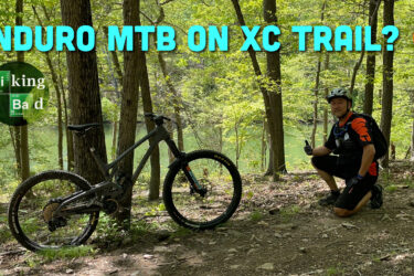 A mountain biker kneeling next to an enduro mountain bike on a cross-country trail, surrounded by lush green trees. The background features a glimpse of a body of water. The image includes text questioning the compatibility of enduro bikes on XC trails. Ten Mile Creek mountain bike trail.