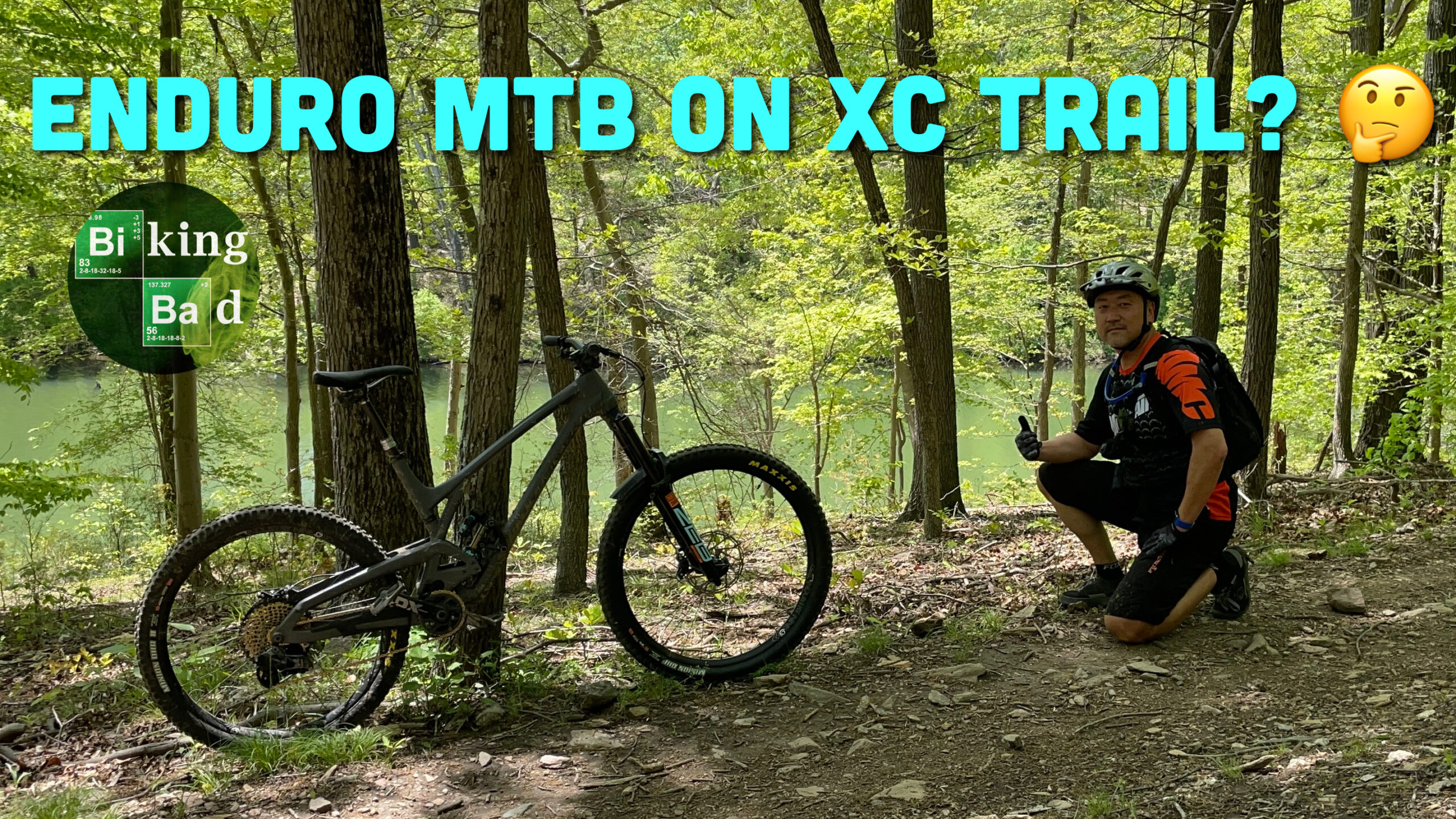A mountain biker kneeling next to an enduro mountain bike on a cross-country trail, surrounded by lush green trees. The background features a glimpse of a body of water. The image includes text questioning the compatibility of enduro bikes on XC trails. Ten Mile Creek mountain bike trail.