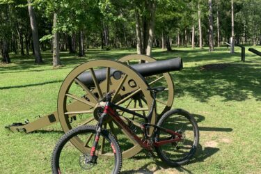 A mountain bike leaning against a green cannon in a park, surrounded by tall trees and grass under bright sunlight. Blakeley State Park mountain bike trail.