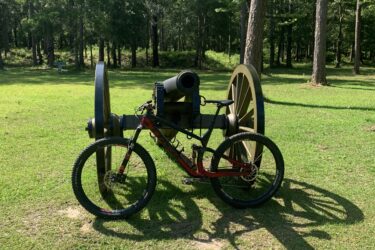 A red mountain bike is positioned next to a historical cannon on a grassy area surrounded by trees. The cannon, featuring large wooden wheels and a black barrel, is placed on a green lawn, which is dotted with sunlight filtering through the leaves above. Blakeley State Park mountain bike trail.