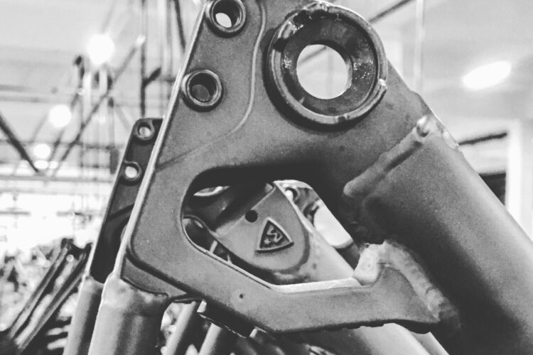 Close-up of a metal bicycle frame component, showcasing intricate details like holes and welds, in a black and white setting. The background features blurred outlines of additional frames hanging in a workshop environment.