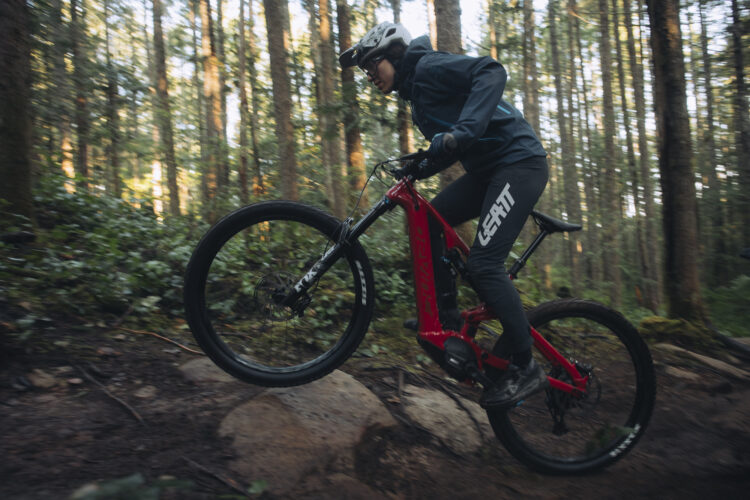 A cyclist in a blue jacket and black pants maneuvers an electric mountain bike over rocky terrain in a forested area. The background features tall trees and greenery, capturing a moment of dynamic movement as the front wheel lifts off the ground.
