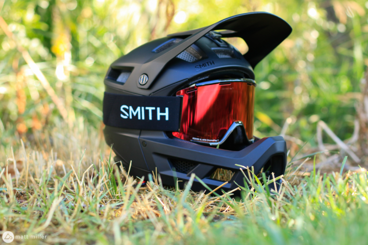 A black mountain bike helmet with a visor and red goggles resting on grass, surrounded by greenery. The helmet features a branded strap with the name "SMITH" prominently displayed.