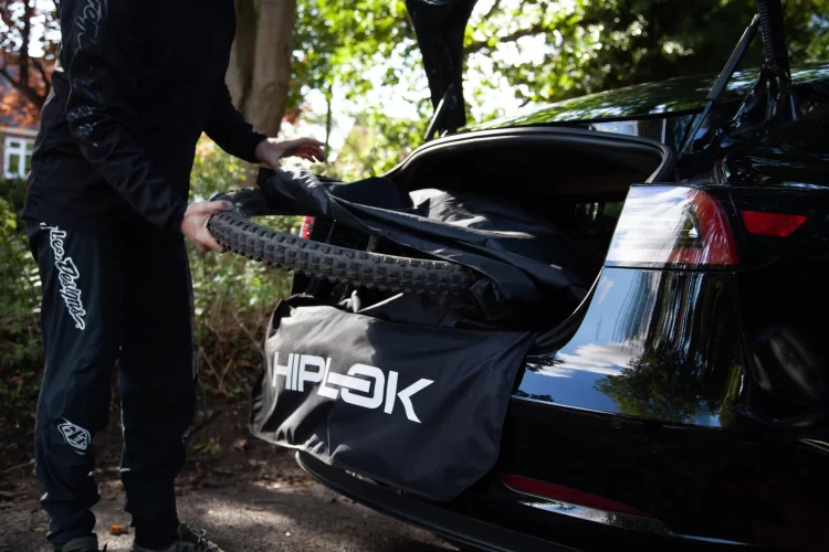 A person is loading a mountain bike tire into the trunk of a black car, with a black storage bag labeled "HIPLOK" partially visible. The scene is set in a park-like environment with trees in the background. The individual is dressed in athletic wear.