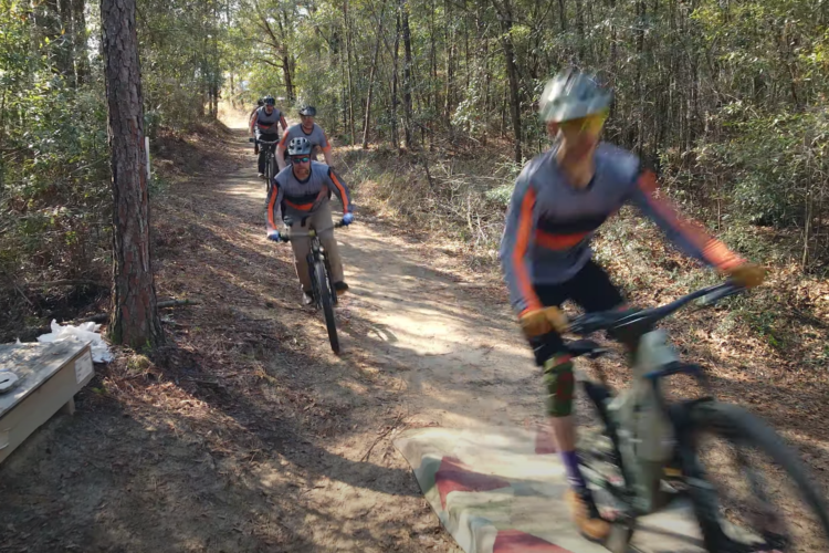 A group of four mountain bikers riding on a dirt trail through a forested area. One biker is in the foreground, leaning to navigate a small jump or ramp, while the others follow closely behind. The setting is sunny, with trees lining the path, and the ground is covered in leaves and dirt.