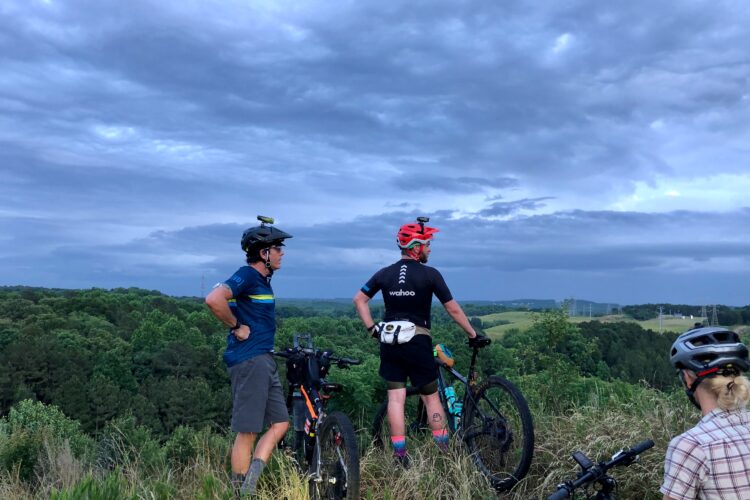 Three mountain bikers are standing on a hillside, overlooking a lush green landscape under a cloudy sky. One biker, wearing a blue shirt and helmet, has his hands on his hips while another, clad in a black shirt with a logo and a red helmet, stands next to his bike. The third biker, wearing a light plaid shirt and a helmet, is slightly turned away from the camera. Bikes are positioned beside them, and the scene captures a moment of appreciation for nature and the outdoors.