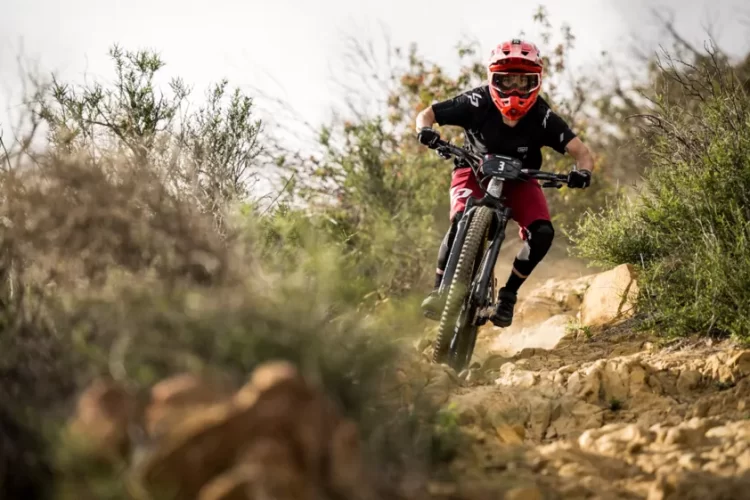A cyclist wearing a red helmet and goggles rides a mountain bike down a rocky trail surrounded by vegetation. The clothing includes a black jersey with red accents and padded shorts. The rider is focused on navigating the challenging terrain, showcasing the excitement of mountain biking.