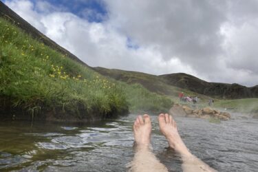 Relaxing feet submerged in warm water, surrounded by lush greenery and yellow wildflowers, with mist rising in the background. Scenic hills and cloudy sky are visible, along with distant figures enjoying the natural setting. Reykjadalur-Nesjavellir mountain bike trail.