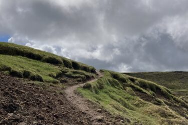 Alt text: A dirt path winds up a grassy hillside under a cloudy sky, leading toward a higher elevation in a natural landscape. Reykjadalur-Nesjavellir mountain bike trail.