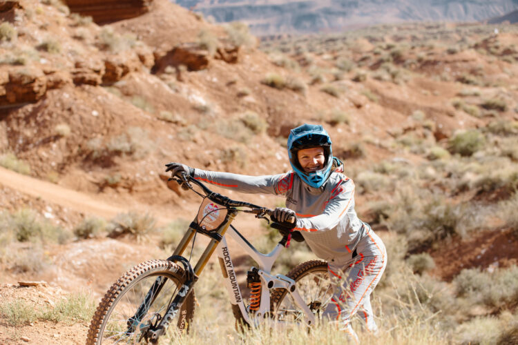 A mountain biker in protective gear smiles while leaning on their bike in a rocky, desert landscape. The bike features a prominent brand logo, and the surrounding terrain is dry with sparse vegetation.