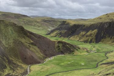 A panoramic view of a lush green valley surrounded by steep, rocky hills under a cloudy sky. A winding stream flows through the valley, and patches of grass and vegetation are visible on the hillsides. The landscape has a mix of earthy tones, with dark rocks contrasting against the vibrant green grass. Reykjadalur-Nesjavellir mountain bike trail.