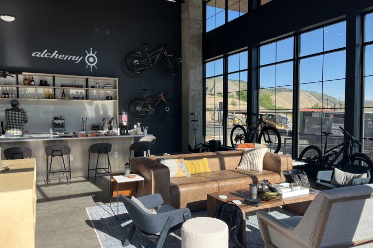 A modern, spacious interior of a café or communal space featuring a stylish bar area with stools and a wall displaying bicycles. The room is furnished with a leather sofa, several armchairs, and tables, offering a cozy atmosphere with large windows that provide ample natural light and a view of the outdoors. A person in a plaid shirt is seen working behind the bar.