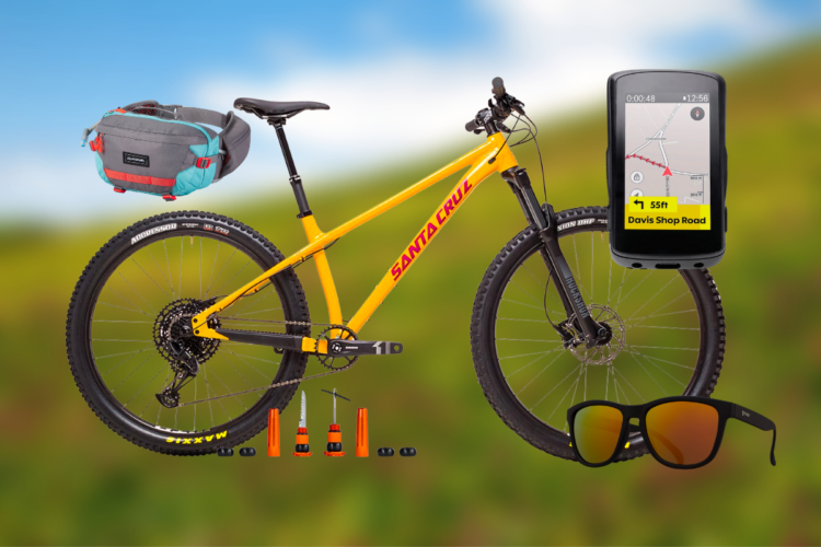 A vibrant yellow mountain bike with the brand name "Santa Cruz" prominently displayed, accompanied by a gray and teal waist bag, a GPS device showing a map, several bike repair tools, and a pair of stylish sunglasses. The background features a blurred natural landscape with green hills and a blue sky.