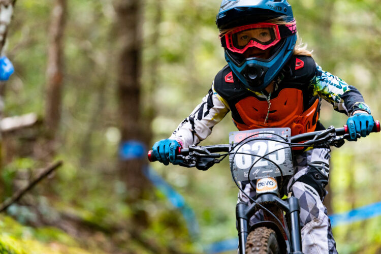A young mountain biker wearing protective gear, including a helmet and goggles, navigates a wooded trail. The athlete is focused and in motion, with a race number displayed on the bike. Trees and blue markers line the trail in the background.