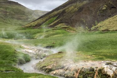 A serene landscape featuring a steaming hot spring flowing through lush green grass, surrounded by rolling hills and rocky terrain under a cloudy sky. Reykjadalur-Nesjavellir mountain bike trail.