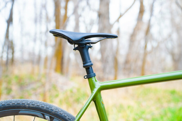 Close-up view of a green bicycle with a black seat, showcasing the seat post and surrounding natural environment with blurred trees in the background.