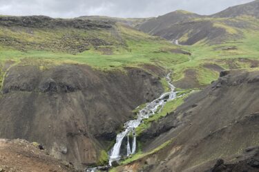 A scenic landscape featuring a series of cascading waterfalls flowing through rocky terrain, surrounded by green hills and gray overcast skies. The image captures the natural beauty and ruggedness of the area, with a mixture of vegetation and barren land. Reykjadalur-Nesjavellir mountain bike trail.