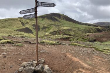 A weathered signpost stands at a trail junction, indicating various hiking paths in a lush, green landscape. The sign points to Reykjadalur (1.0 km), Hveragerði (7.3 km), and other trails, surrounded by rolling hills and a cloudy sky. Reykjadalur-Nesjavellir mountain bike trail.