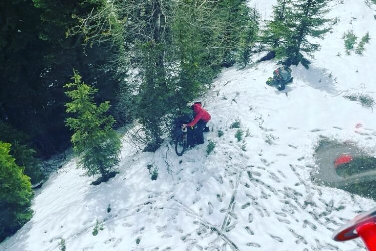A snowy landscape with green trees, featuring two individuals engaged in outdoor activities. One person in a red jacket is near a bicycle, while the other is further down the slope, surrounded by snow. Footprints are visible in the snow, indicating recent foot traffic.