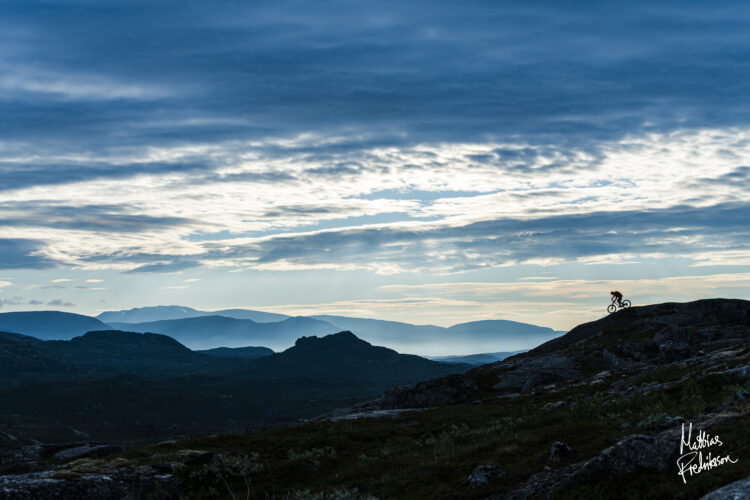 A silhouette of a mountain biker riding across rocky terrain under a dramatic sky filled with clouds. In the background, rolling hills fade into the distance. The soft light of sunset casts a serene atmosphere over the landscape.