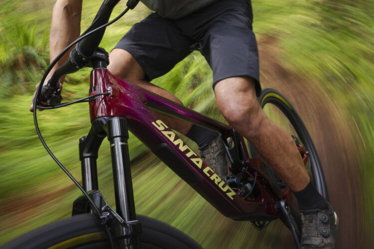 A mountain biker riding a purple Santa Cruz bike on a dirt trail surrounded by lush greenery, with motion blur conveying speed and excitement.