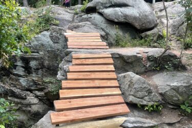 A wooden bridge made of slats spans between large rocks in a wooded area. In the background, there is a cabin deck partially visible, surrounded by tall trees. A person can be seen walking up the rocky path toward the deck. Battle Park mountain bike trail.