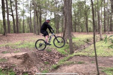 A cyclist in a black outfit and helmet is jumping off a small dirt ramp in a wooded area, surrounded by green trees and fallen leaves. The biker's front wheel is lifted off the ground as he navigates the terrain. Battle Park mountain bike trail.
