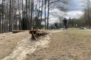 A mountain biker is seen jumping off a wooden ramp on a dirt trail, with several other cyclists resting nearby on a grassy area surrounded by trees. The sky is cloudy, indicating overcast weather. Battle Park mountain bike trail.