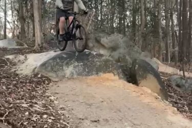 A mountain biker jumping off a rock on a dirt trail surrounded by trees and fallen leaves. Battle Park mountain bike trail.