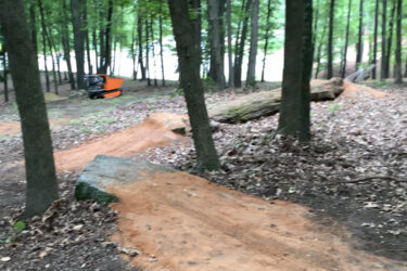 A winding dirt path through a forested area, surrounded by tall trees and scattered leaves. A large, partially visible log lies on the ground, with an orange piece of equipment in the background near a clearing. The scene appears serene and natural, showcasing a setting suitable for outdoor activities. Battle Park mountain bike trail.