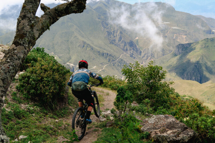 A mountain biker descends a narrow trail surrounded by lush greenery, with dramatic mountainous terrain and misty clouds in the background. The rider is wearing a helmet and a blue jersey, navigating the rocky path on a mountain bike.