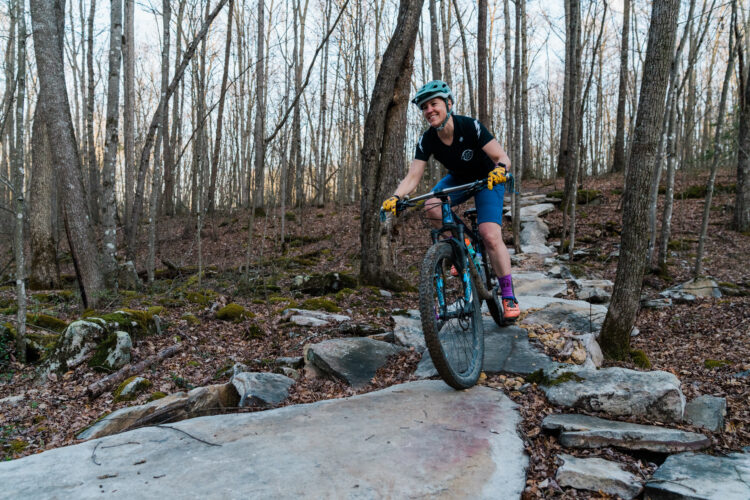 A person riding a mountain bike on a rocky trail in a wooded area. The cyclist is wearing a helmet, a black shirt, and blue shorts, with colorful socks and gloves. The forest features bare trees, patches of moss, and scattered rocks, creating a natural biking environment.