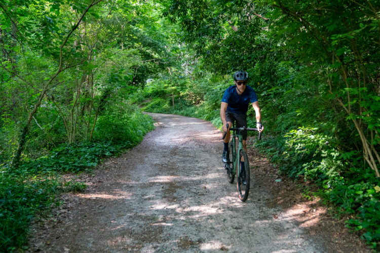 A cyclist riding on a dirt path surrounded by lush green trees and foliage. The path winds through a wooded area, indicating a scenic outdoor biking route. The cyclist is wearing a helmet and athletic clothing, actively enjoying the ride.