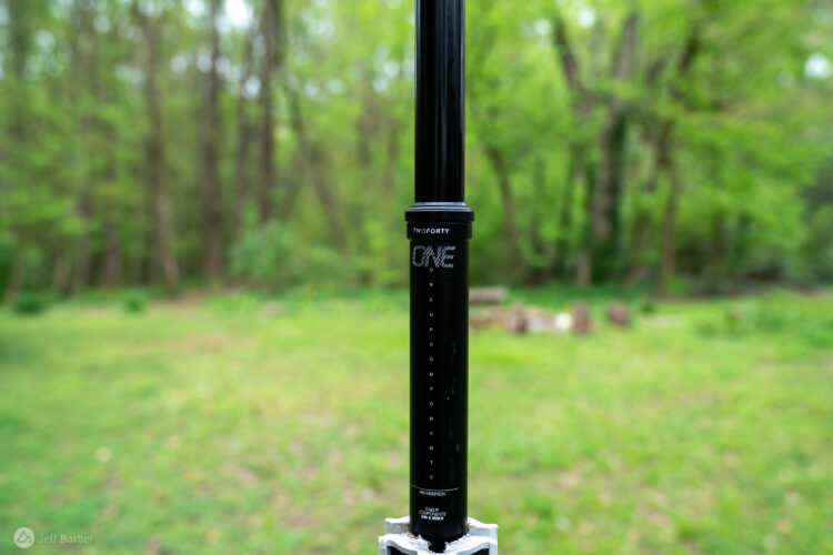 A close-up image of a black bicycle dropper post, featuring the brand name "ONE" and the model "TWOFORTY" printed on it. The background shows a blurred natural landscape with green trees and grass.