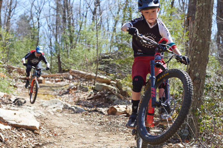 Two young boys are riding mountain bikes on a rocky trail through a wooded area. One boy is performing a jump over a large rock, while the other is visible in the background navigating the path. The scene is bright and sunny, highlighting the natural surroundings and the excitement of mountain biking.